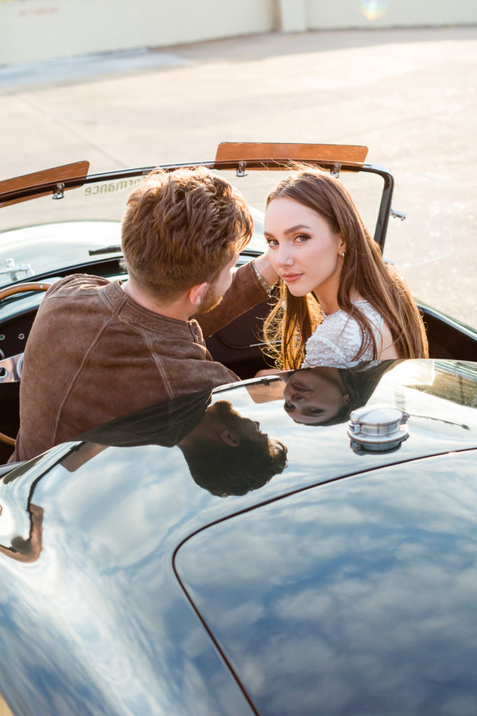 A young couple sitting in a classic car, the bride looking at the camera, the groom looking at her. 