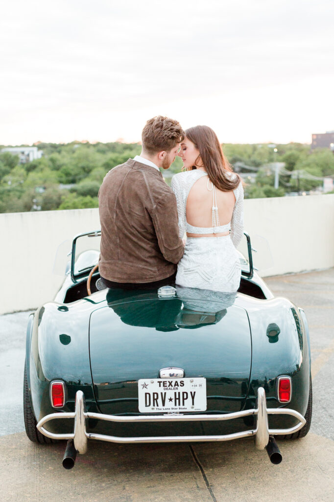 A couple wearing neutral colors about to kiss while sitting on a classic car. 